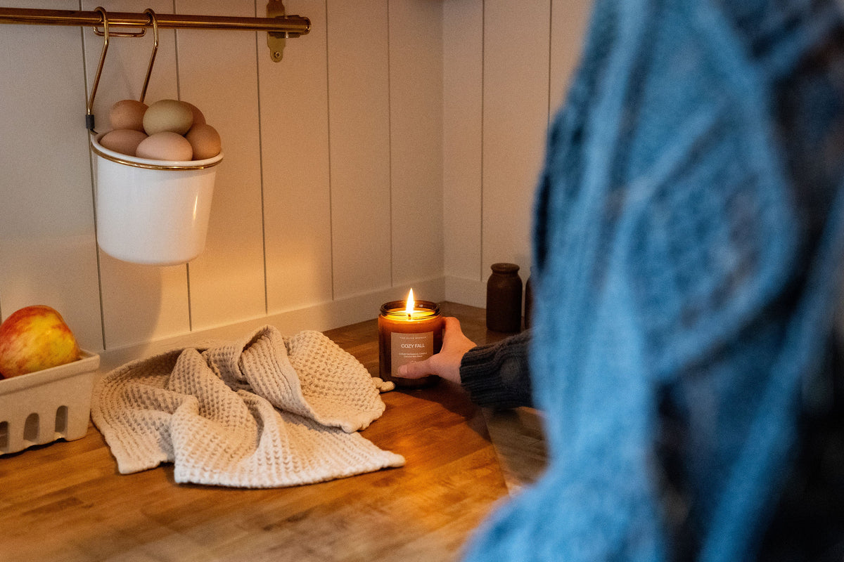 A person in a blue cable-knit sweater holds a lit candle on a wooden kitchen counter, with a waffle-knit towel, hanging eggs, and fruit visible in the background.