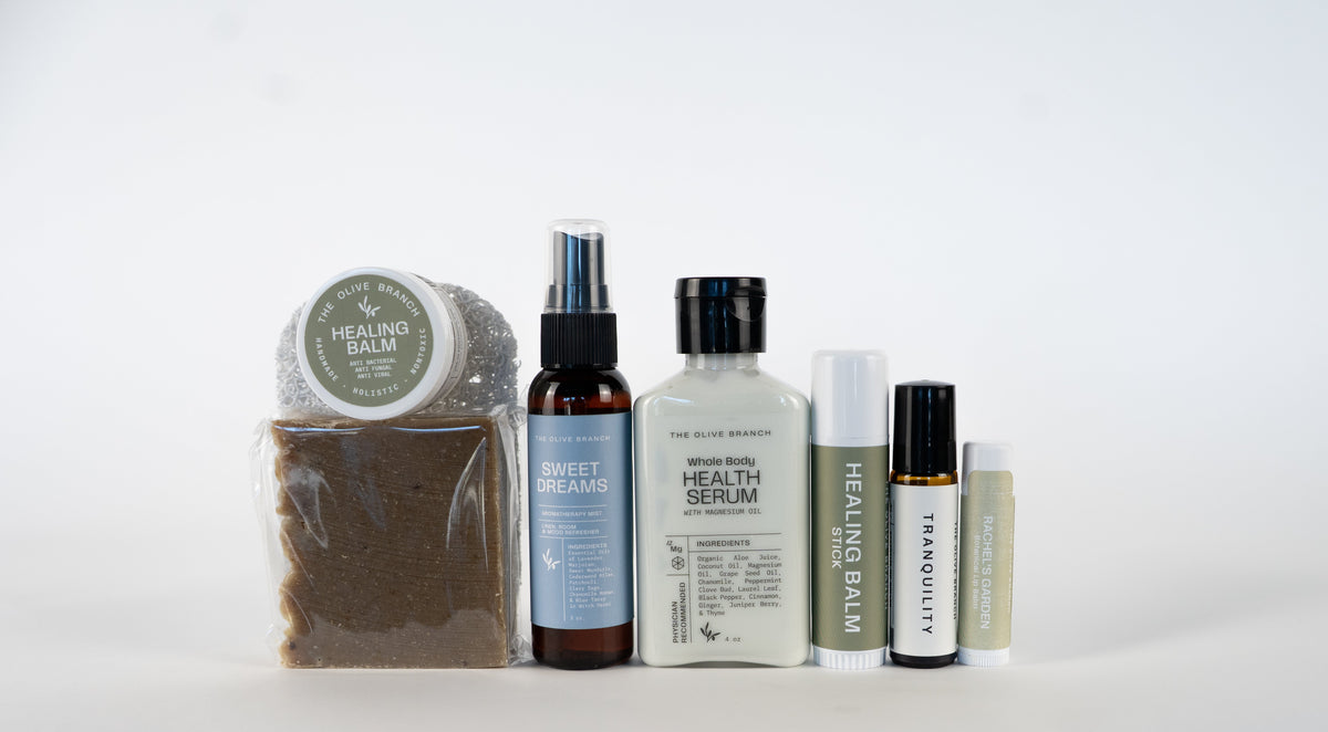 A group of wellness and skincare products arranged on a white surface against a white background, therapeutic botanical soap bar, including healing balm, a Sweet Dream spray, a whole body health serum, and lip balm, tranquility essential oil roller.