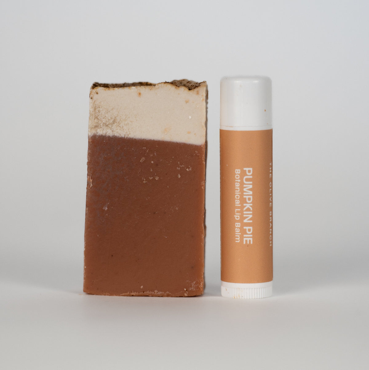 A bar of mini pumpkin pie soap next to a matching lip balm tube with a white cap, both featuring a warm tan label with white text, set against a plain light gray background.
