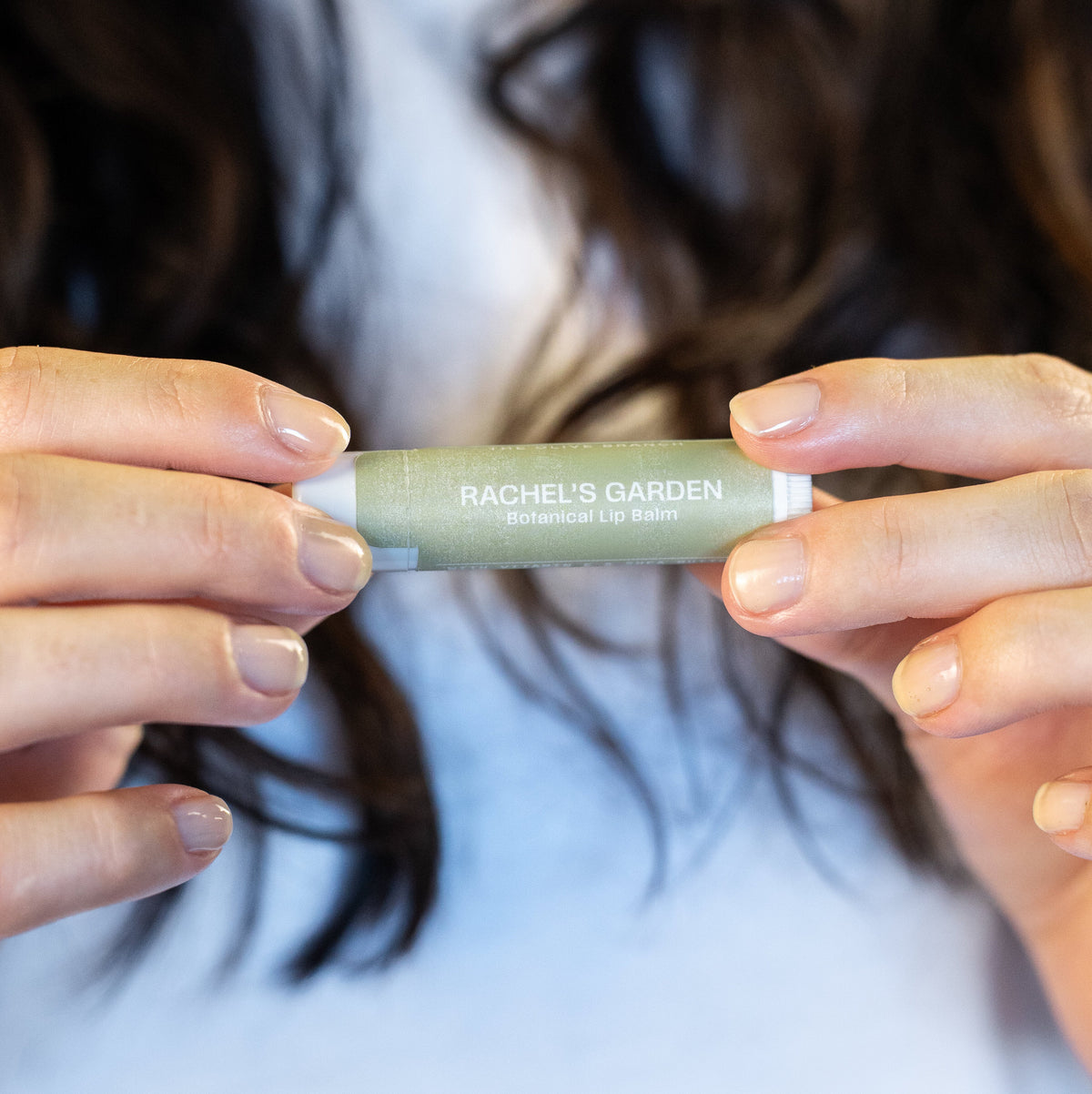 A person holding a slim, light sage green cosmetic stick with white ends. The stick is labeled 'RACHEL'S GARDEN' and 'Sage Green'.