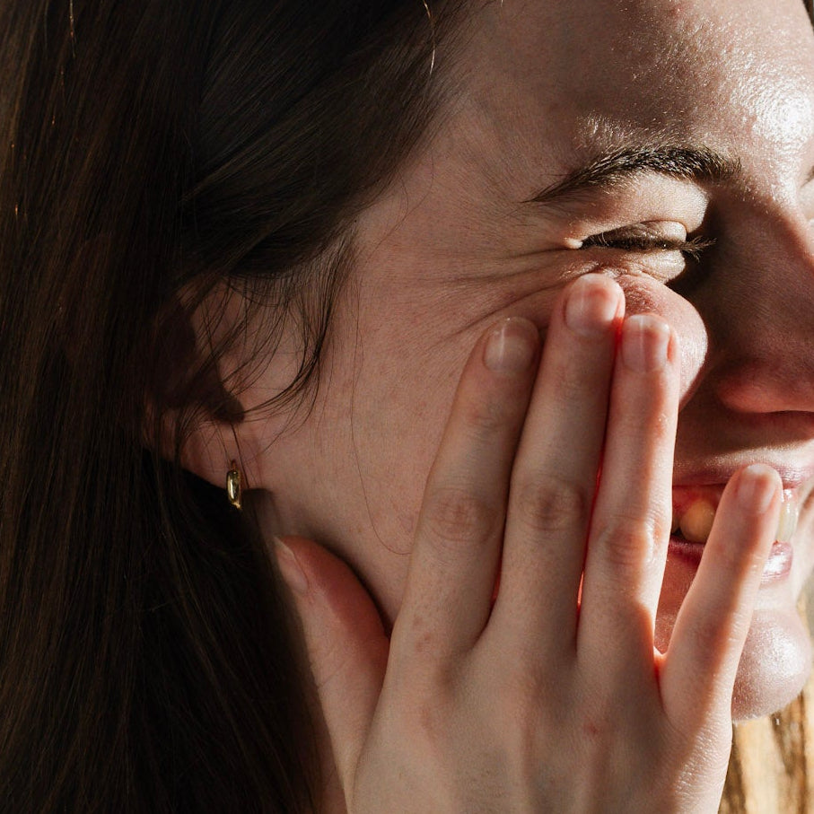 Woman with long hair smiling and holding her hand near her face in a bright room.