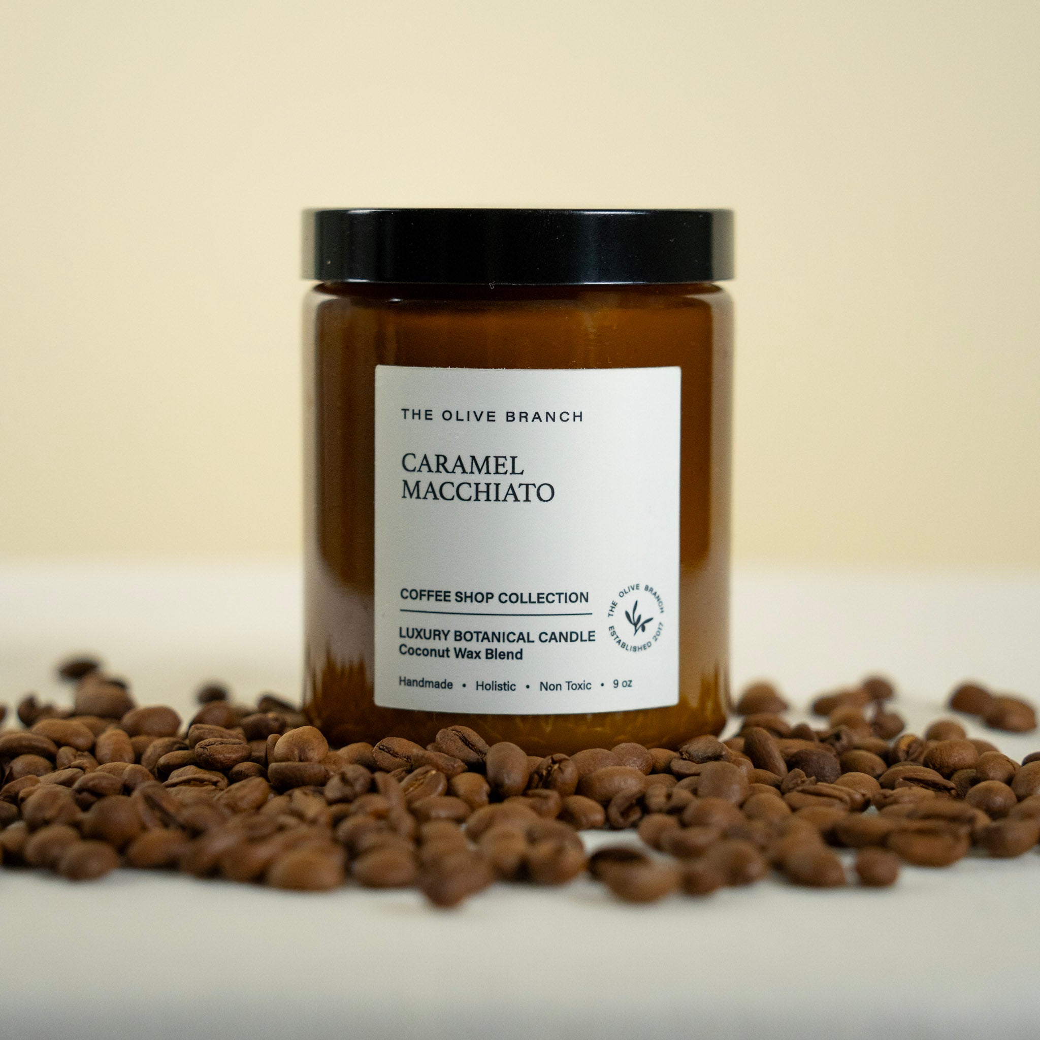 Candle jar labeled 'Caramel Macchiato' from 'The Olive Branch' on a surface with coffee beans.