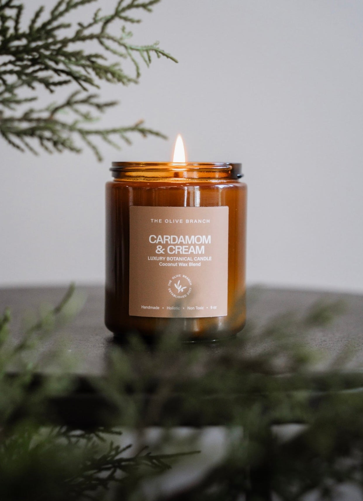 Candle in an amber jar labeled 'Cardamom & Cream' with greenery in the foreground.