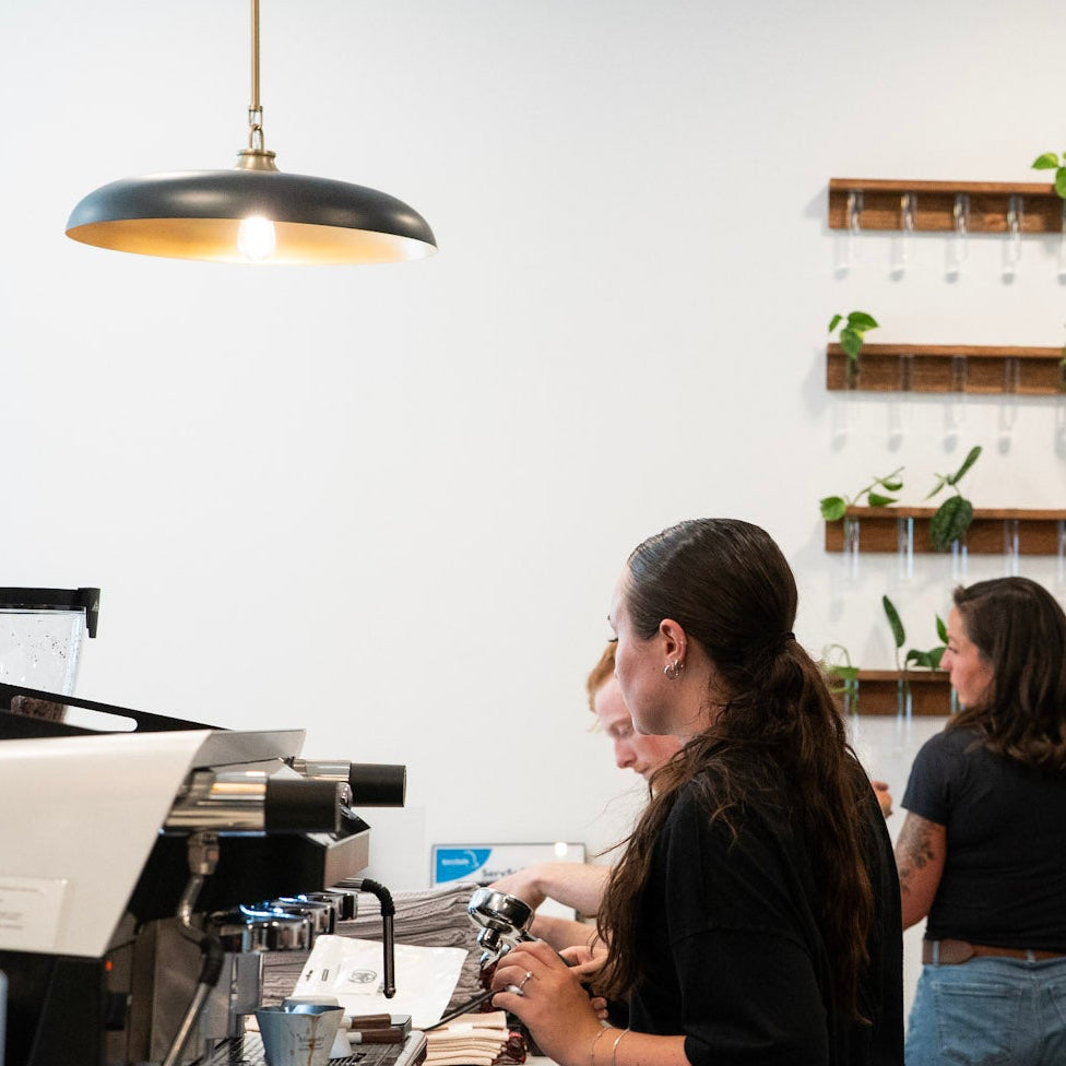 Three women in black t-shirts and jeans working inside a modern cafe or kitchen space with a white wall, wooden floating plant shelves, and a hanging pendant light.