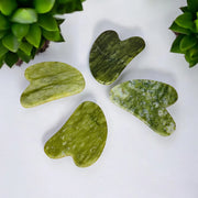 A collection of green jade gua sha stones of various shades, displayed against a white background with some green leaves at the top.