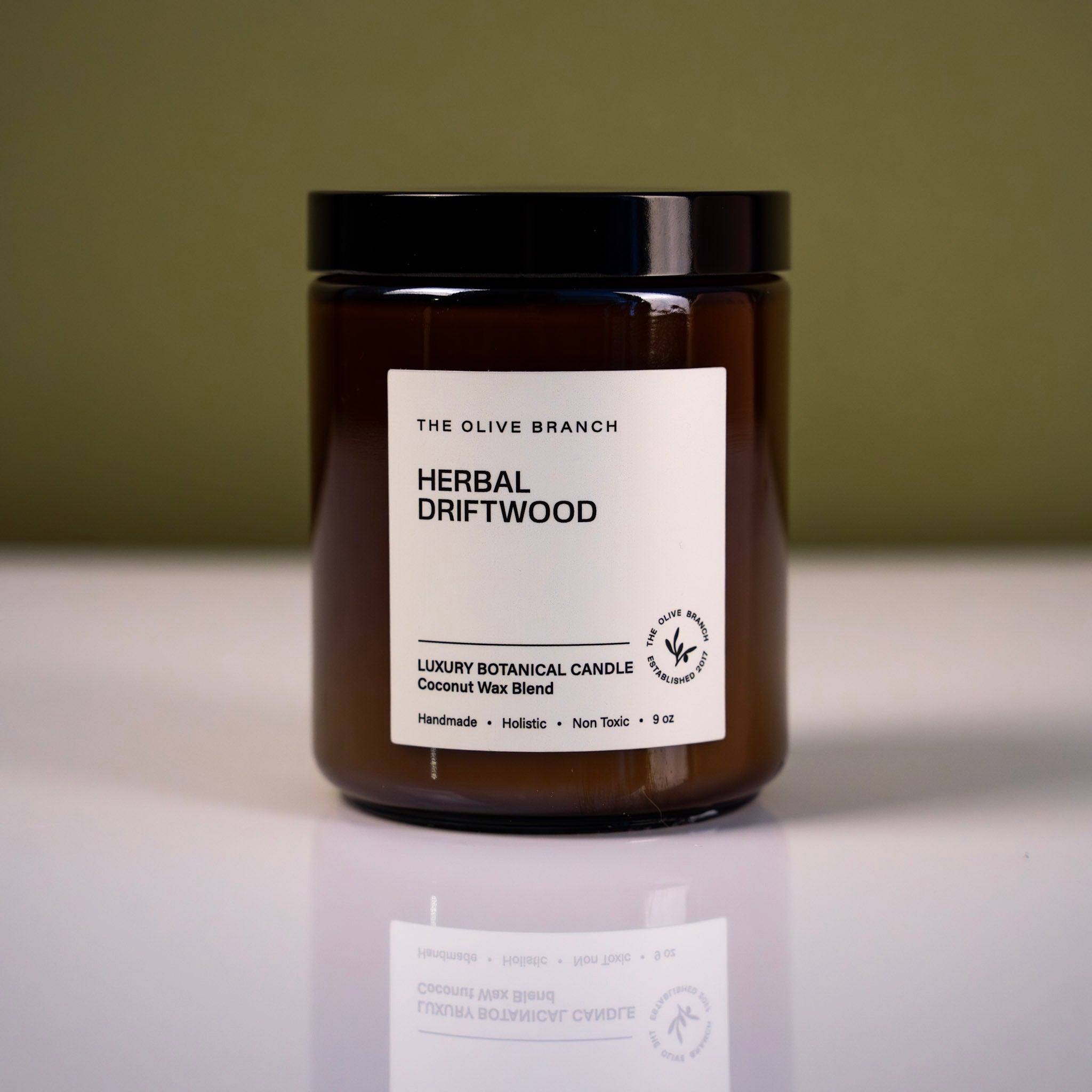 Brown candle jar labeled 'Herbal Driftwood' from 'The Olive Branch' on a reflective surface with a green background.