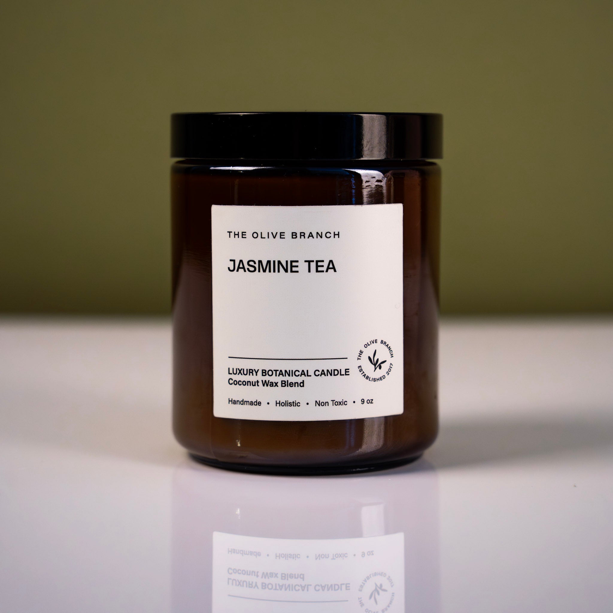Brown candle jar with labeled 'Jasmine tea' on a reflective surface and green background