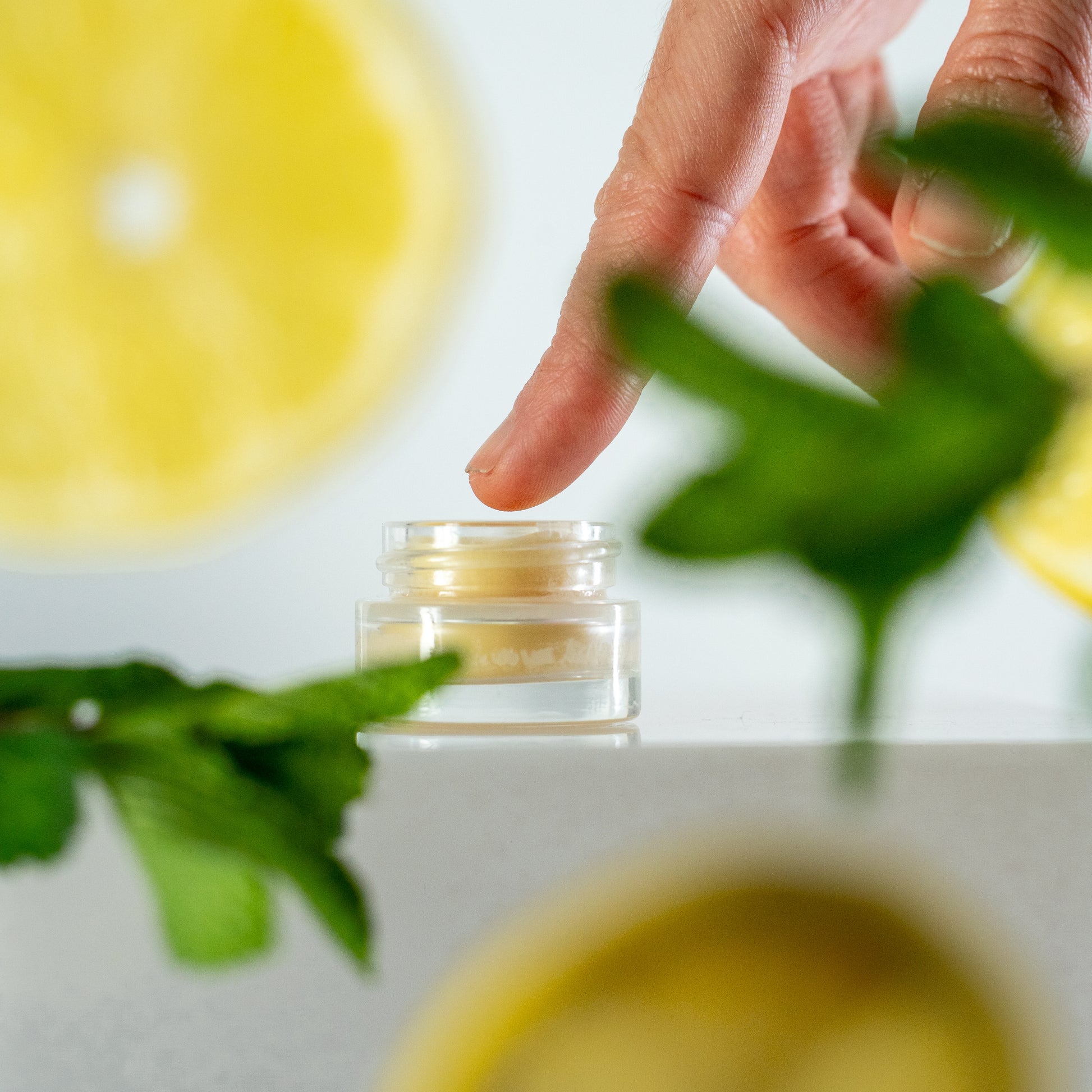 Hand holding a small jar with lip mask surrounded by green leaves on a light background