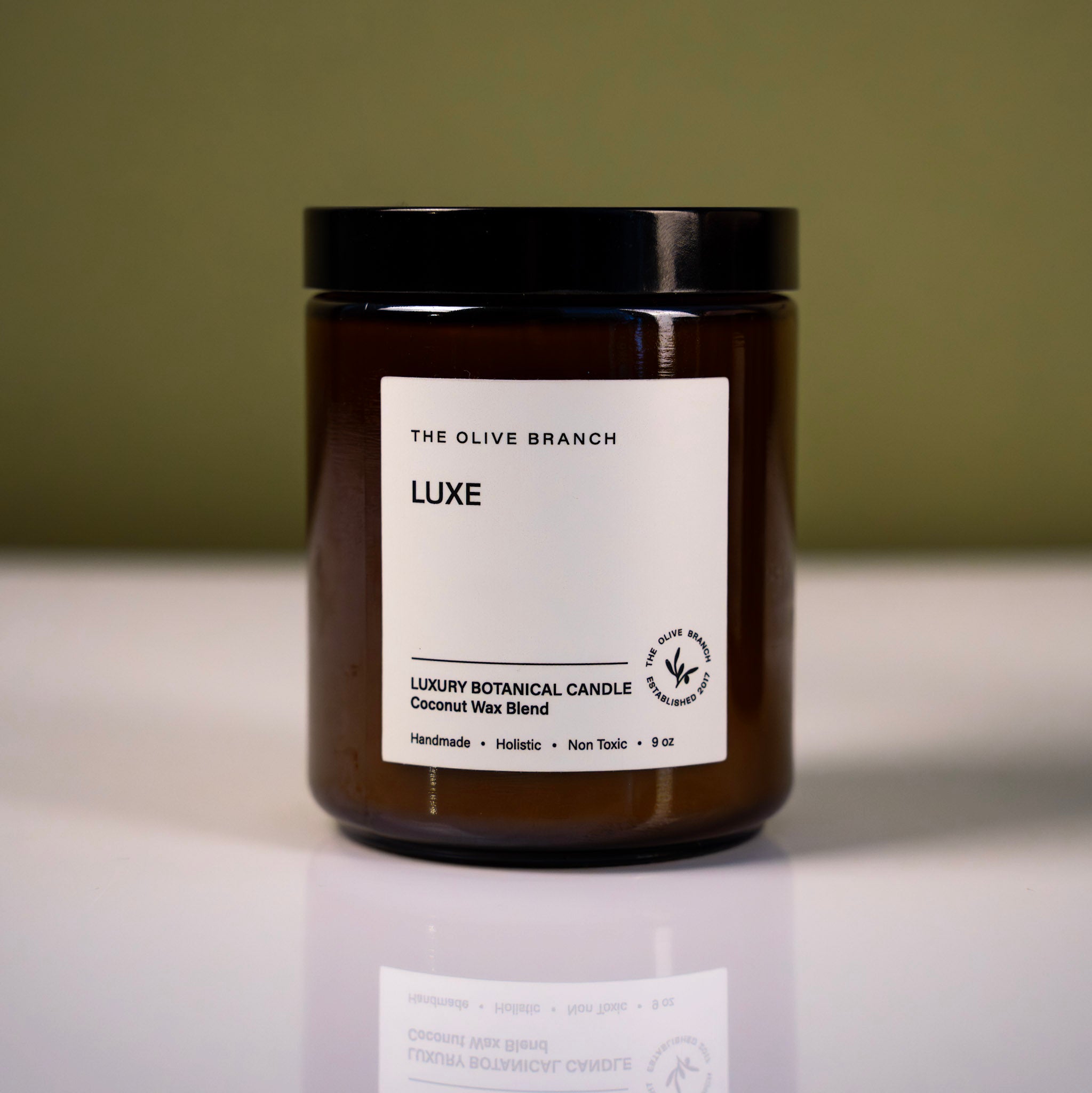 Brown candle jar with white label 'luxe' on a reflective surface and green background
