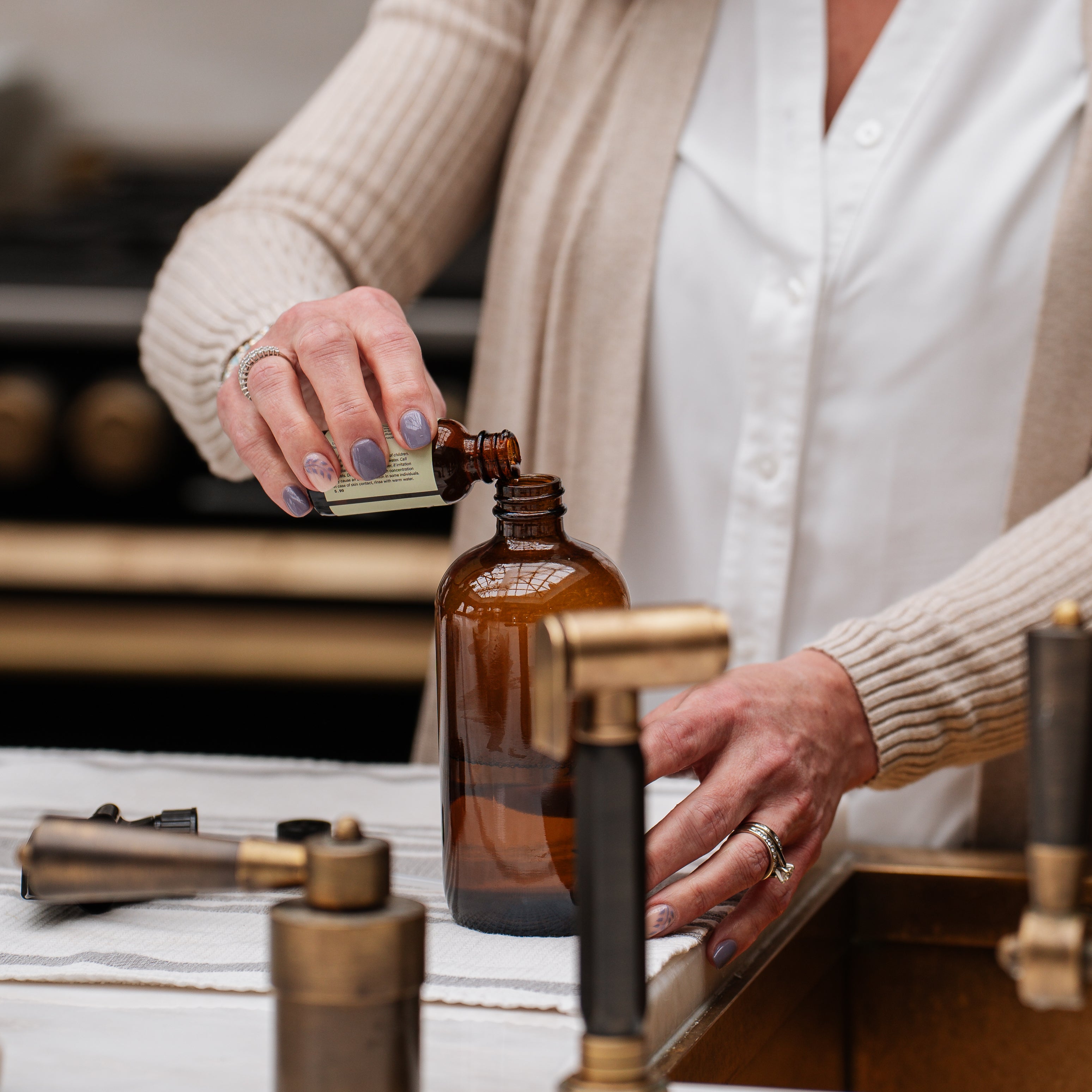 Person pouring multi-surface cleaner concentrate from a small bottle into a larger amber glass bottle on a wooden surface.
