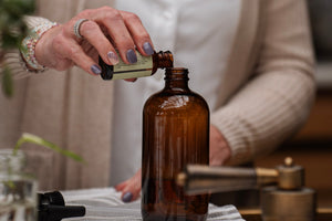Person pouring liquid from a small bottle into a larger brown glass bottle on a table.