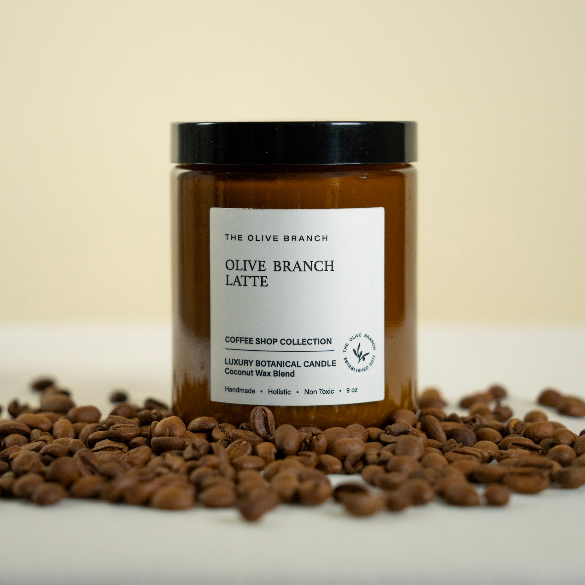Candle jar labeled 'Olive Branch Latte' surrounded by coffee beans on a light background