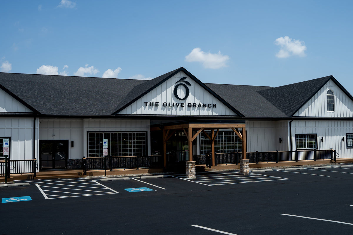 The Olive Branch home and body care store exterior under a blue sky, featuring white siding, a dark gray roof, a wooden covered entryway, and an empty asphalt parking lot in the foreground.