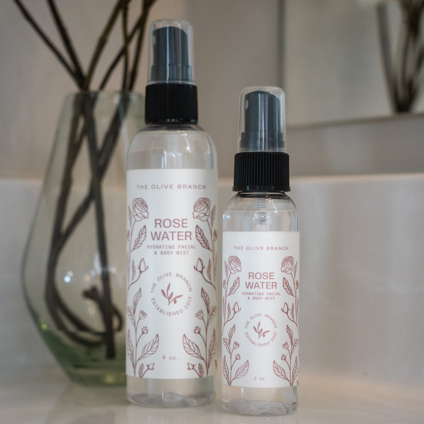 Two bottles of Rose Water spray on a bathroom counter with a vase in the background.