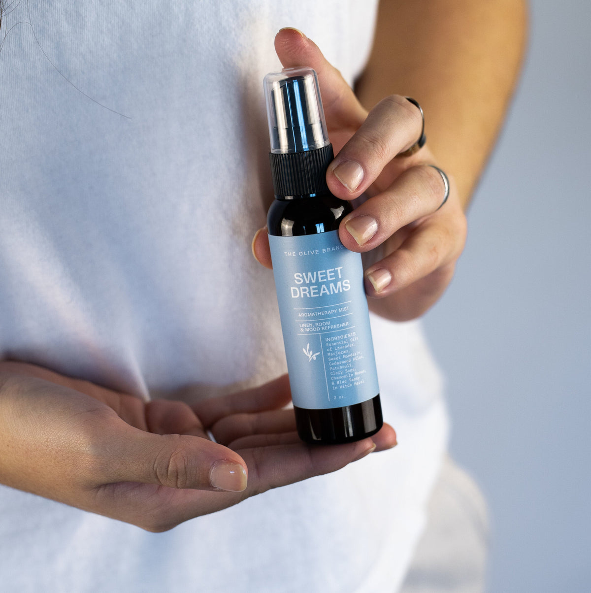 A person in a white t-shirt holds a small dark bottle called sweet dreams with a silver spray top and a light blue label.