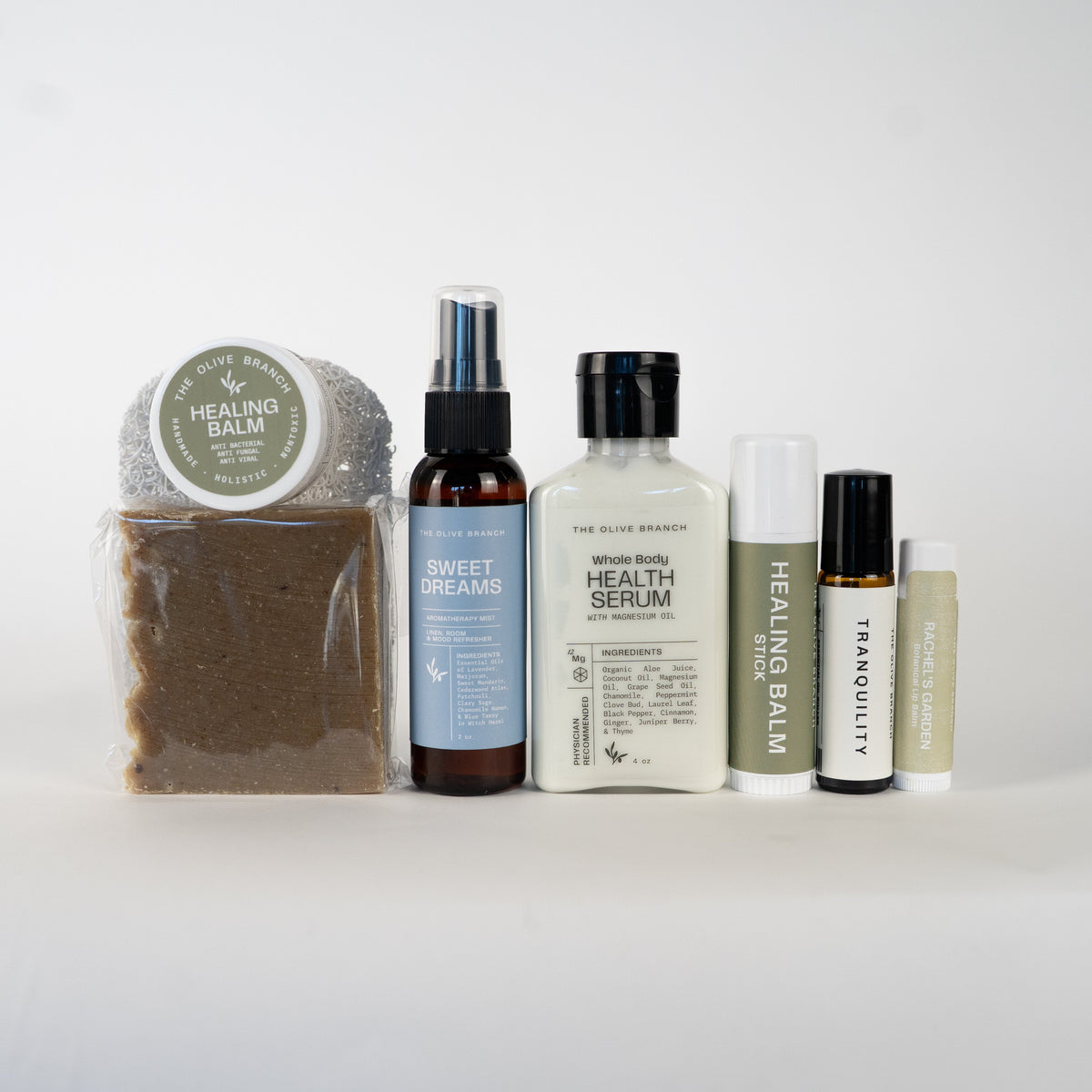 A collection of healing balm products from The Olive Branch arranged on a seamless white background. The set includes a packaged bar of healing balm, a spray bottle, a cream bottle, and two small roller bottles.