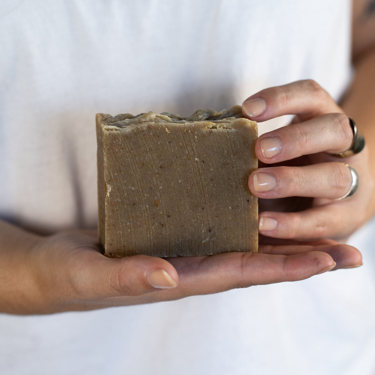 A person in a white shirt holds a rectangular bar of handmade soap in both hands. The soap is a warm brownish-gray color with a rustic, uneven top edge.