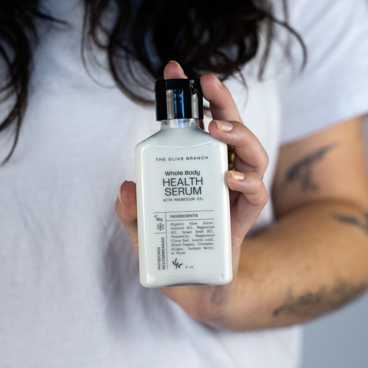 A person in a white t-shirt holds up a white bottle of 'The Olive Branch' Whole Body Health Serum against a neutral gray background.