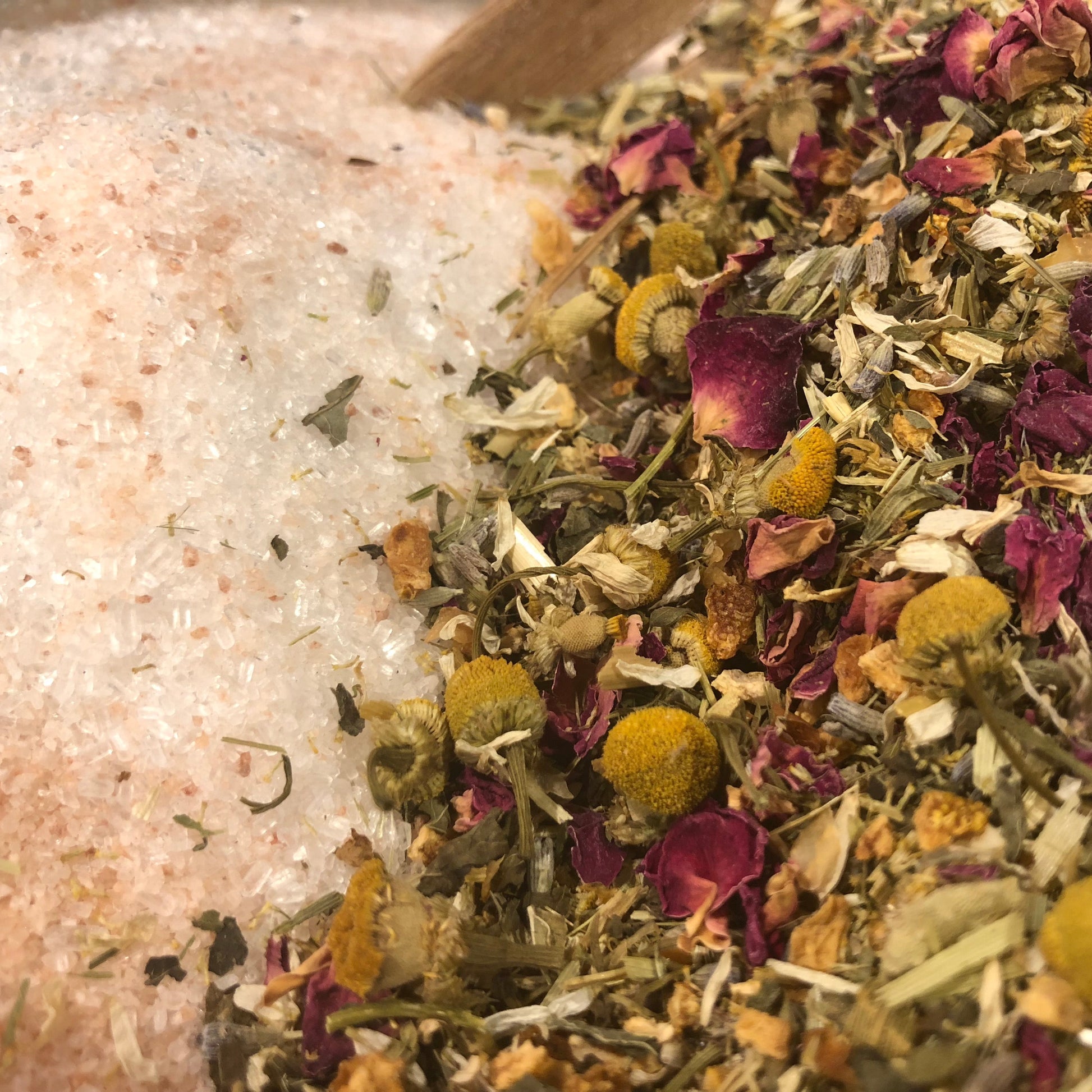 A variety of dried botanicals including chamomile, rose petals, orange peel, oatstraw, lemon balm, and lavender, alongside Epsom salt and Pink Himalayan salt, arranged on a surface.