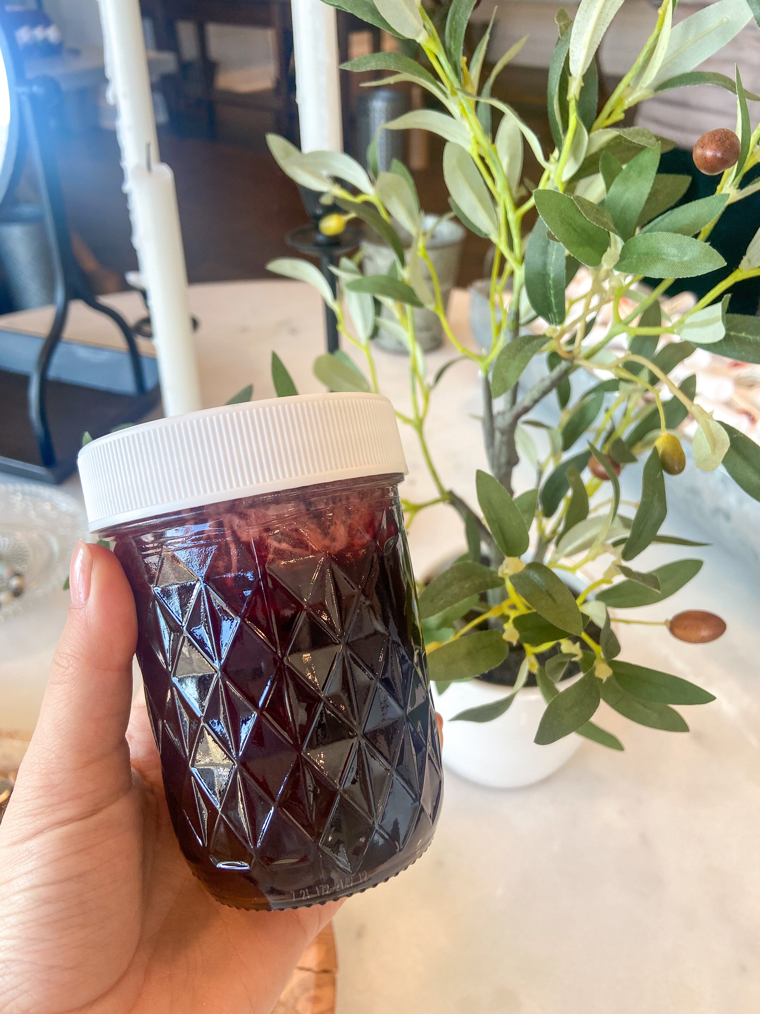 A jar of Hibiscus Honey held in front of a plant, the honey appears dark red.